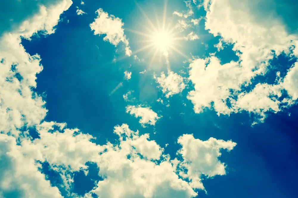 Rays of sunlight shine through fluffy clouds against a vibrant blue sky